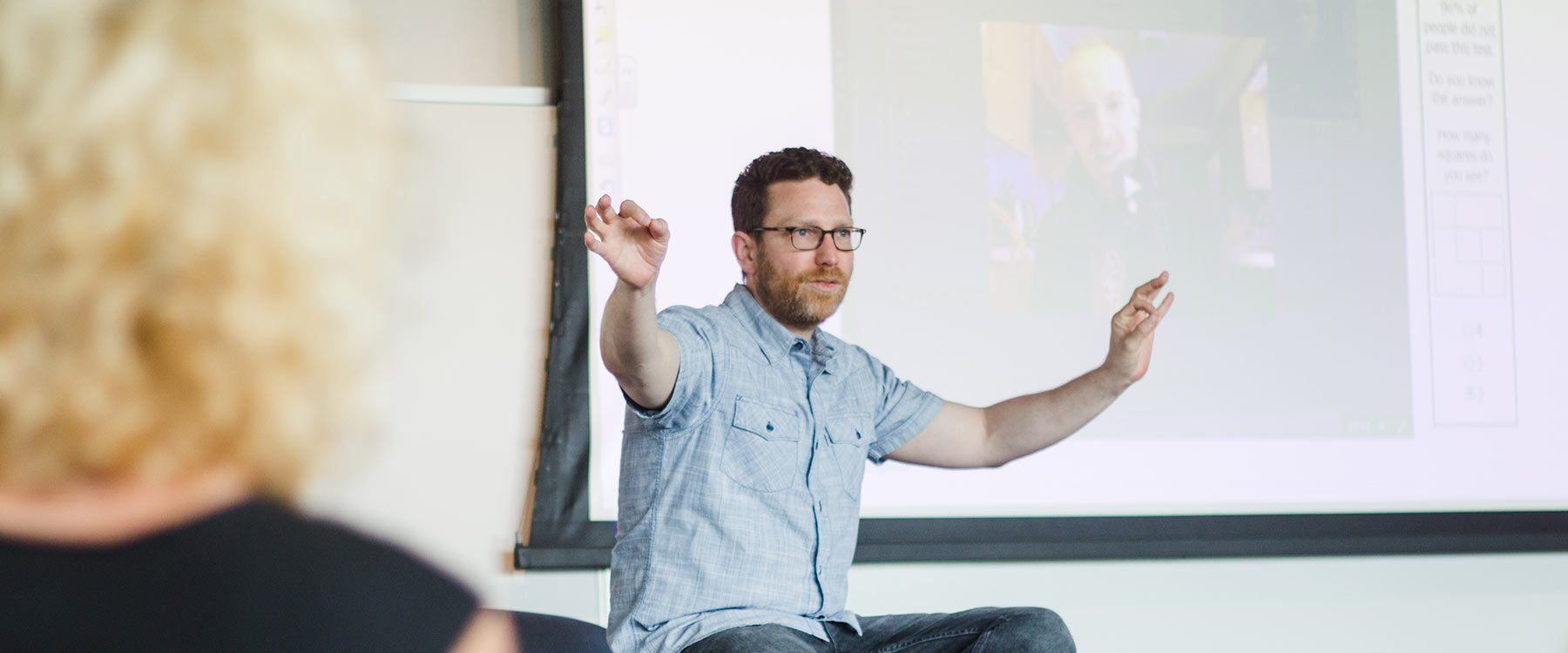 Professor Fred Johnson gestures with both arms in front of a projector screen, presenting an English lecture.