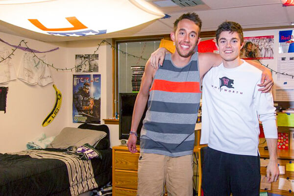 Two male students pose with their arms around the other's shoulders in their dorm room.