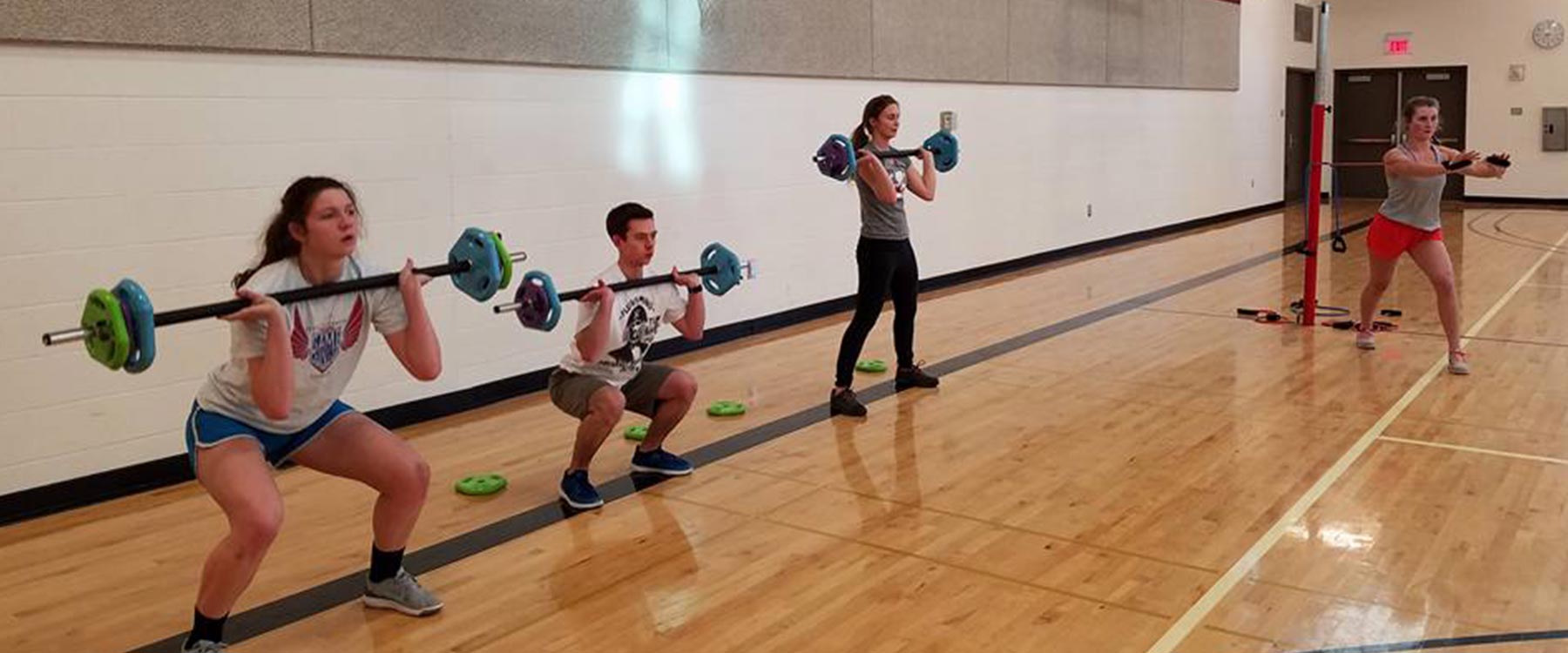 Three students squat with small barbells in a group fitness class.