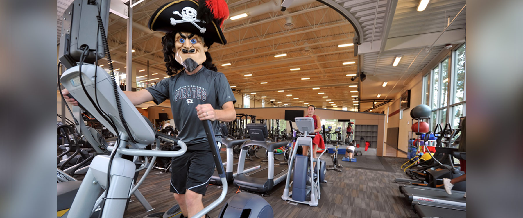 A student wearing the pirate mascot head works out on an elliptical machine.