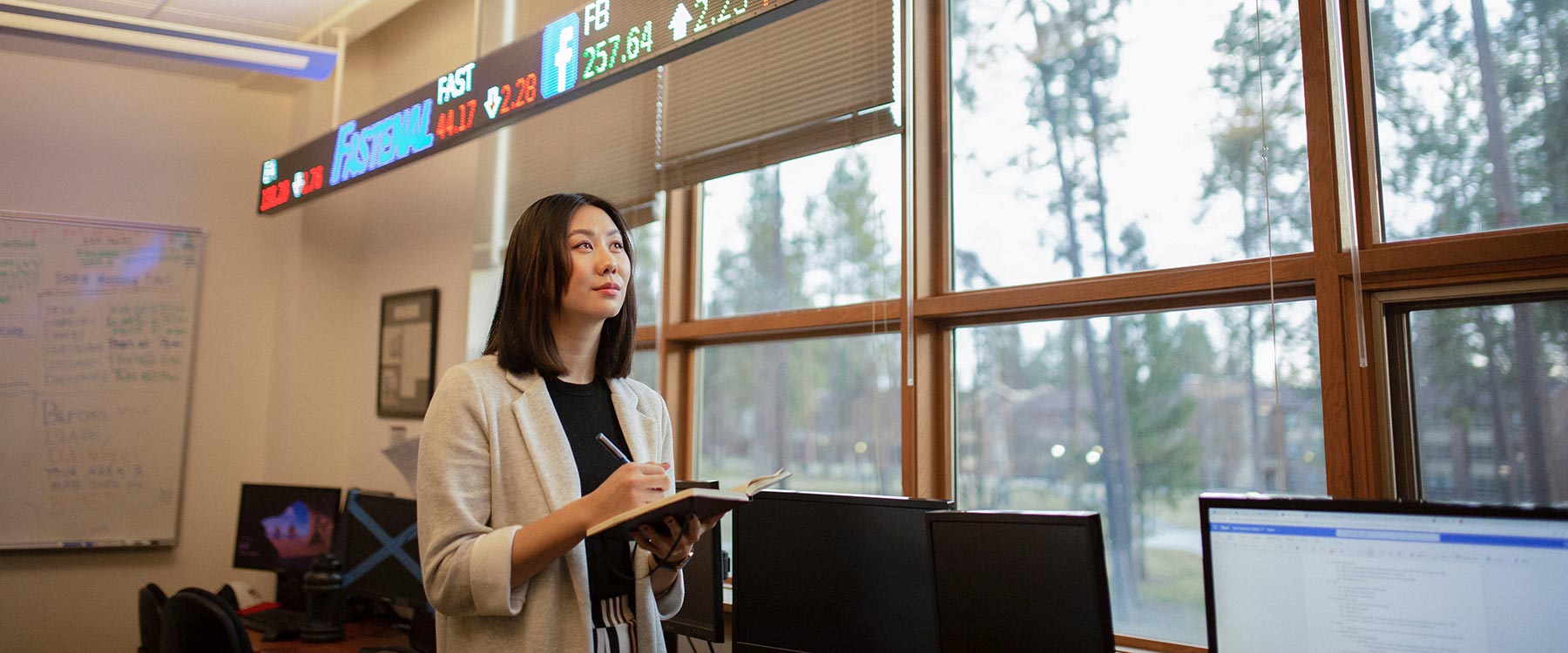 A student takes notes in an office with multiple computer monitors and a stock ticker display on the wall.