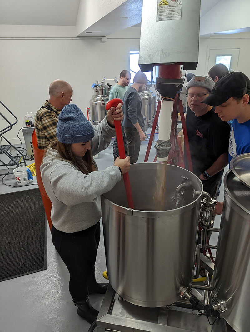 People brewing beer together, one stirring a large vat, others observing and adjusting equipment.