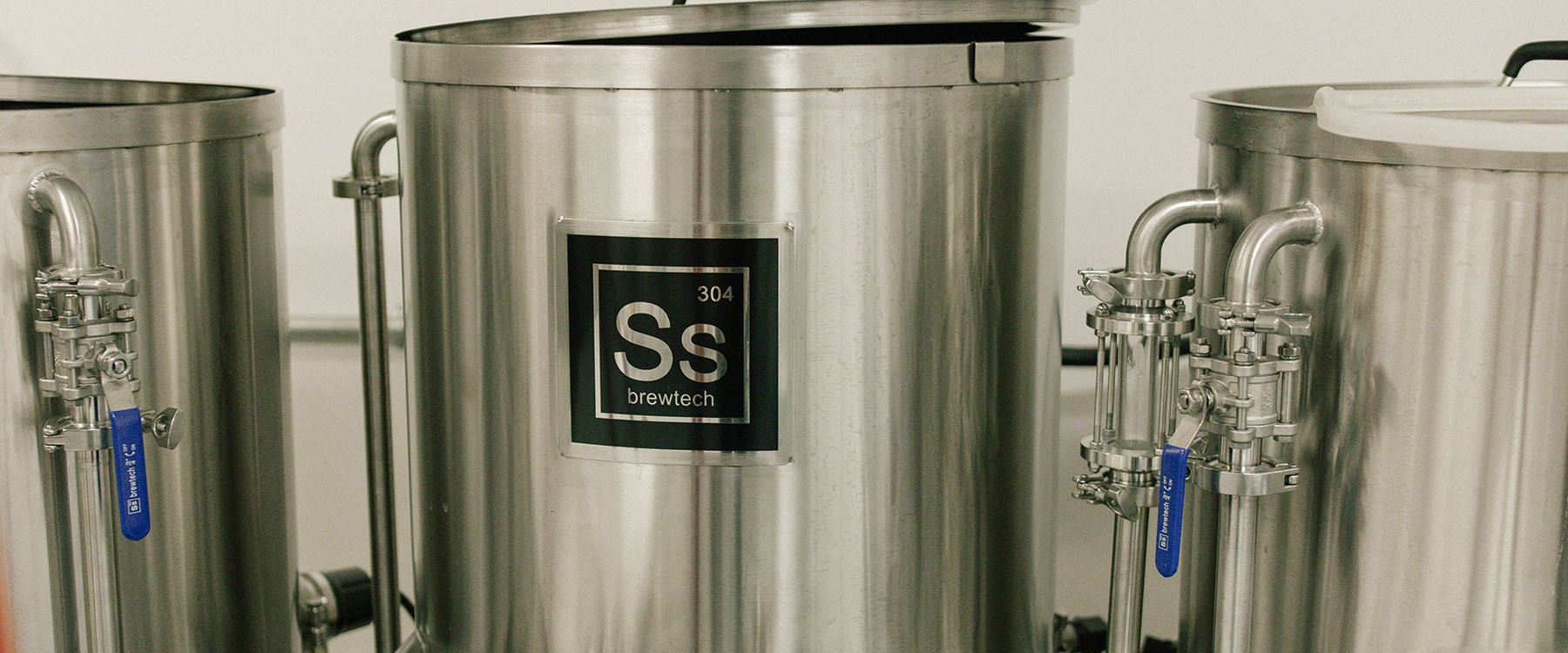 A row of stainless steel brewing tanks with labeled valves and a sign reading 
