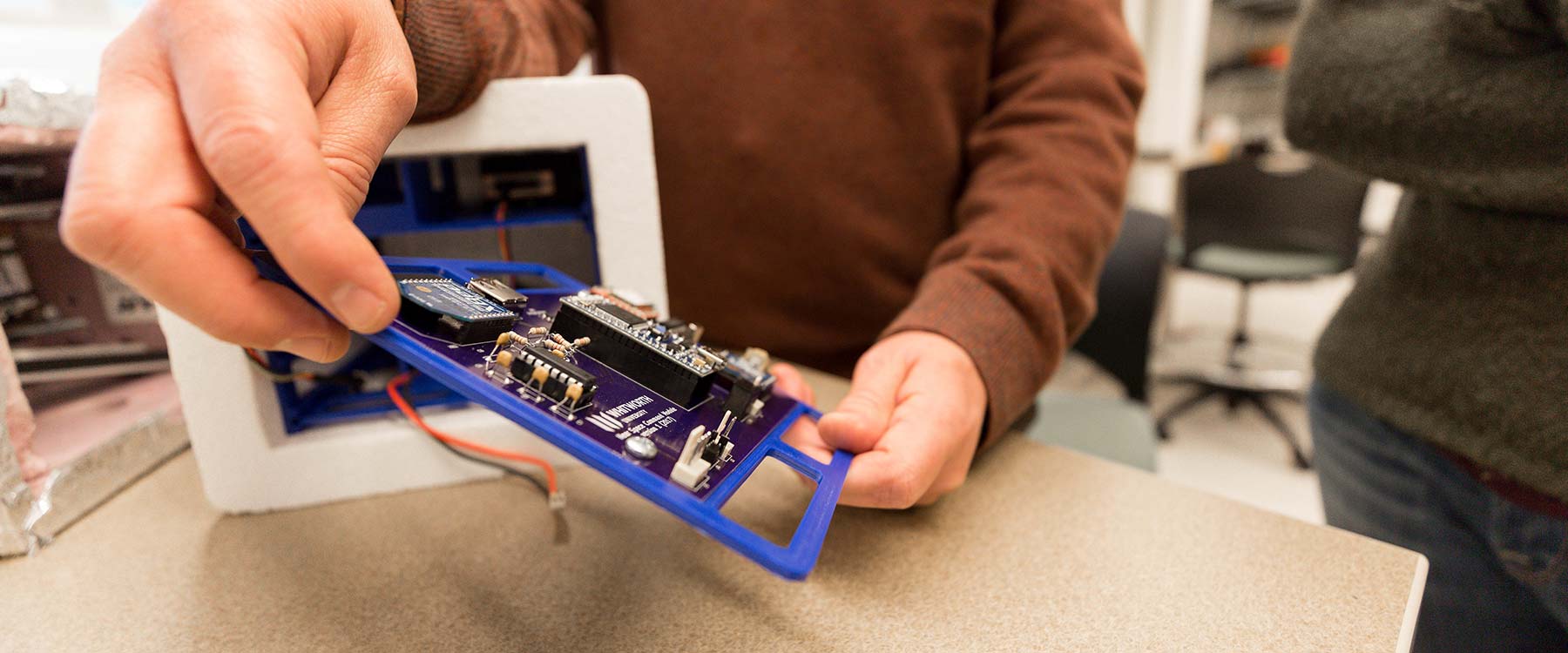 Someone holds a circuit board over a table, tilts it to the side and examines it.