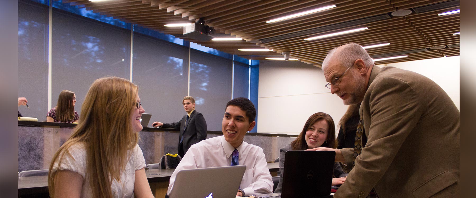 Three students sit in a row behind a table. On the other side, a professor leans on the table and speaks with them.