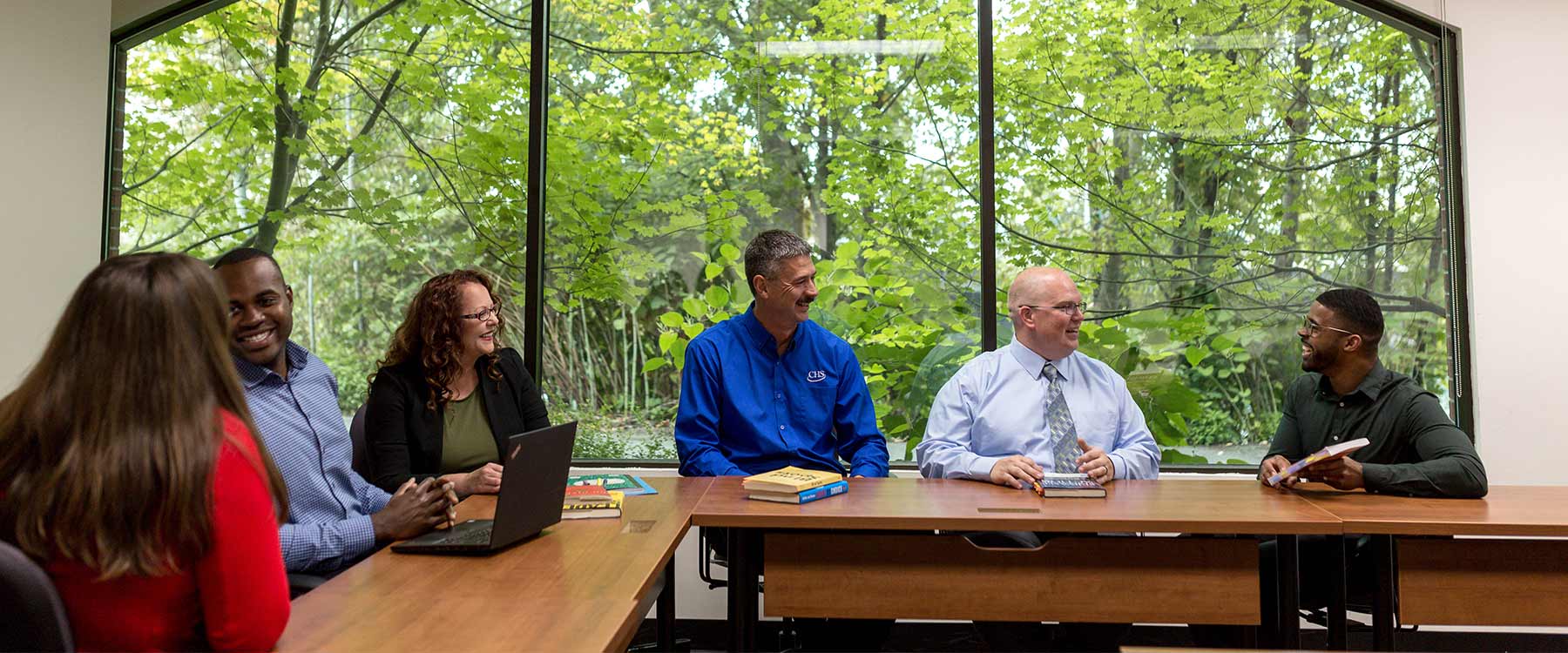 Six adult students sit at a long desk in front of a large window showing green foliage, talking and smiling.