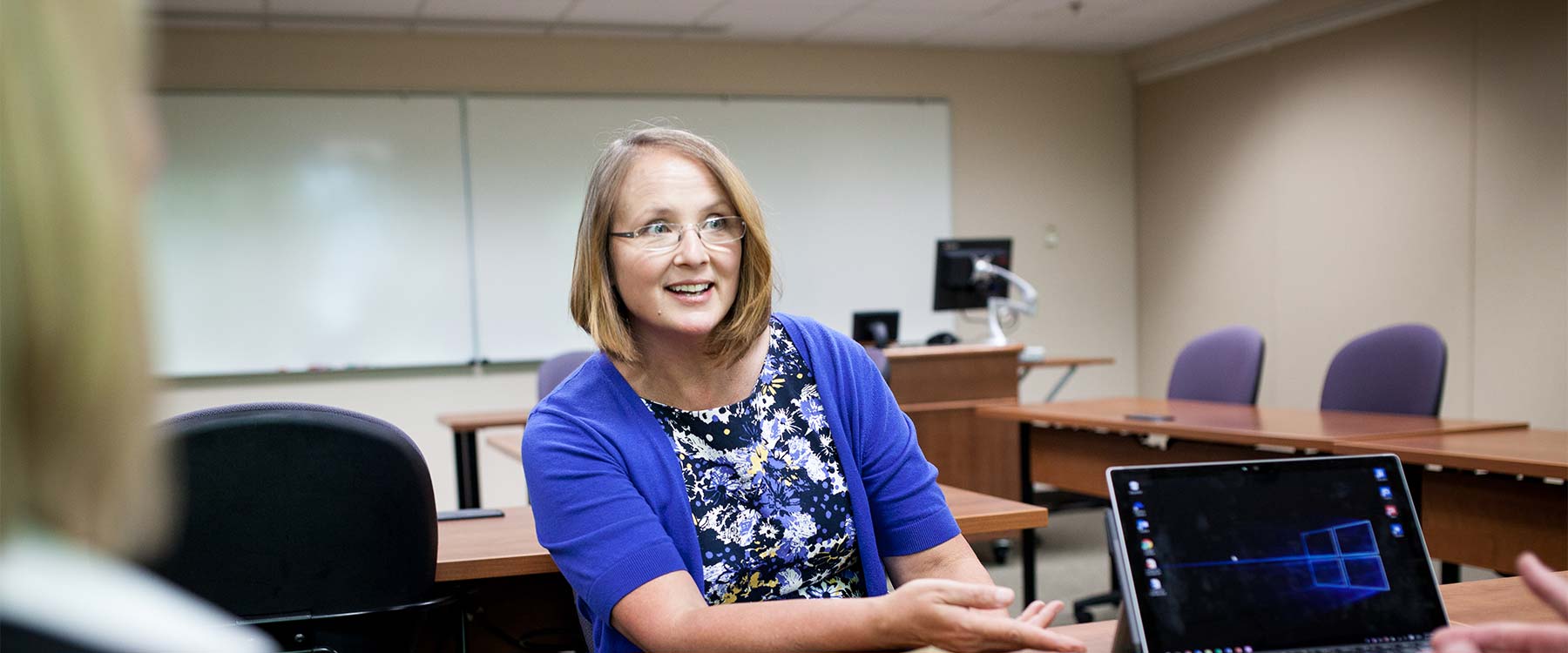 A teacher sits across a desk from two students in a classroom.