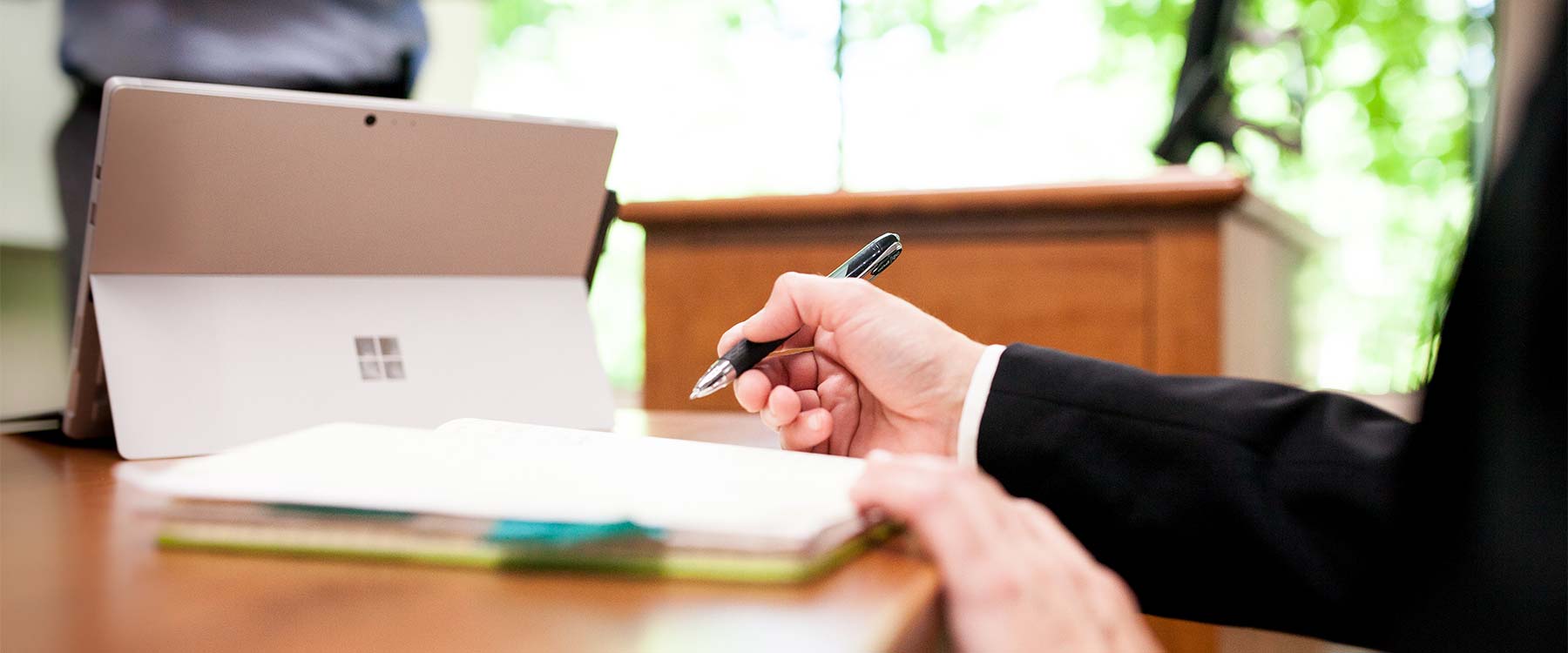 A tablet and notebook sit on a large table. A student holds a pen, poised to write in the notebook.