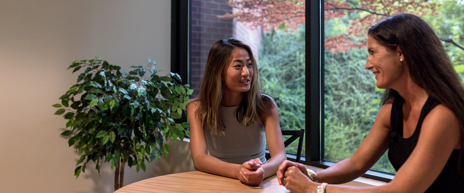 Two women sit at a round table by a window, engaged in conversation, with a plant in the background.