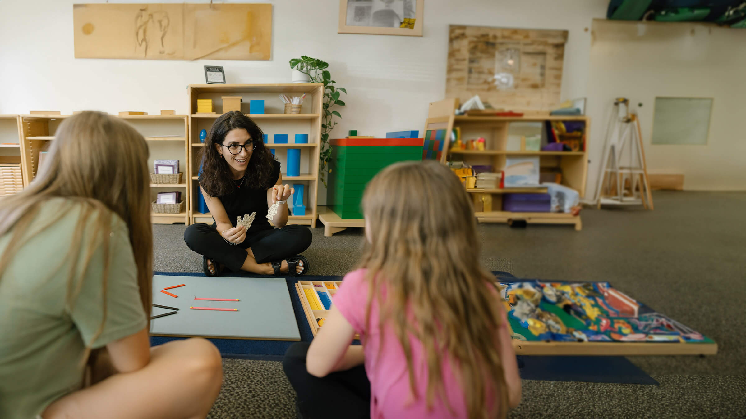 A teacher sits across from two elementary students. They work together building a structure with blocks.