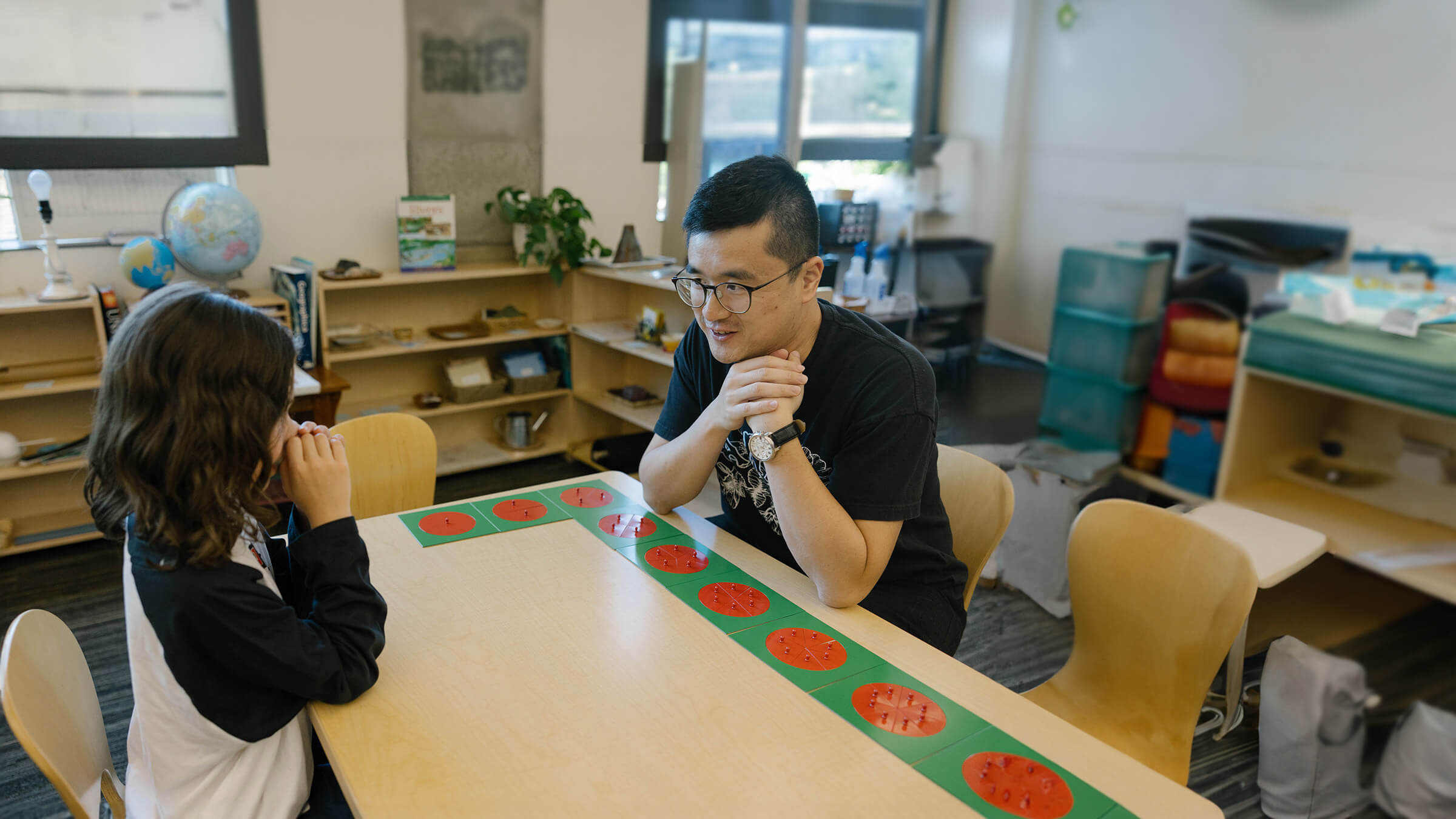 A teacher and a child sit across from each other at a classroom table discussing a project with fractions.