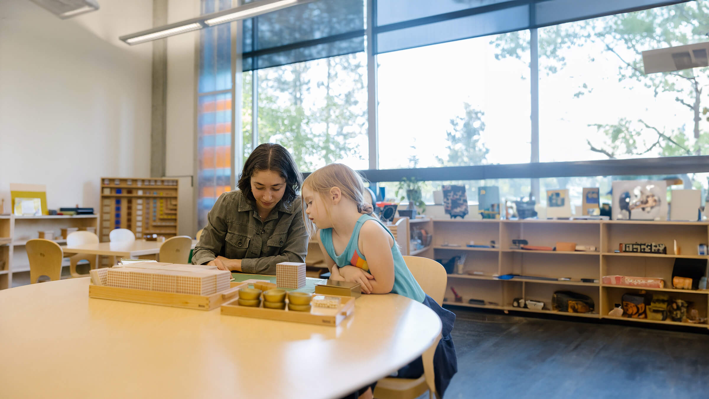 A teacher and child work together at a round table in a well-lit classroom with educational materials.