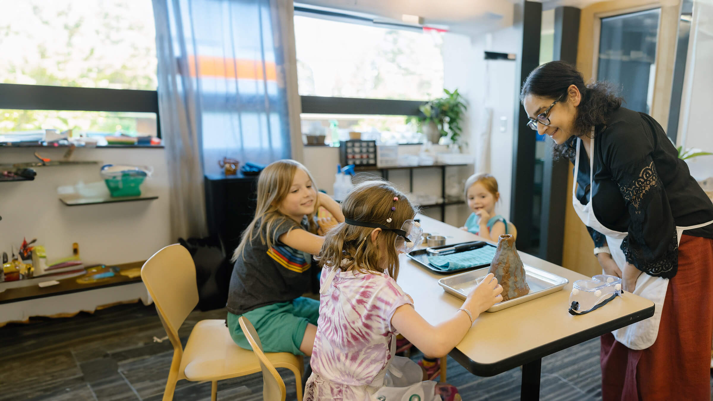 Three children and a teacher in a classroom, engaged in a science experiment with a small volcano model on a table.