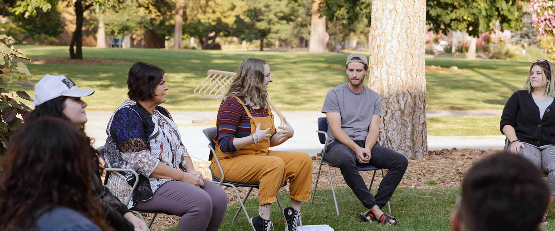 A group of people sit outdoors in a circle, engaging in conversation under the shade of trees.
