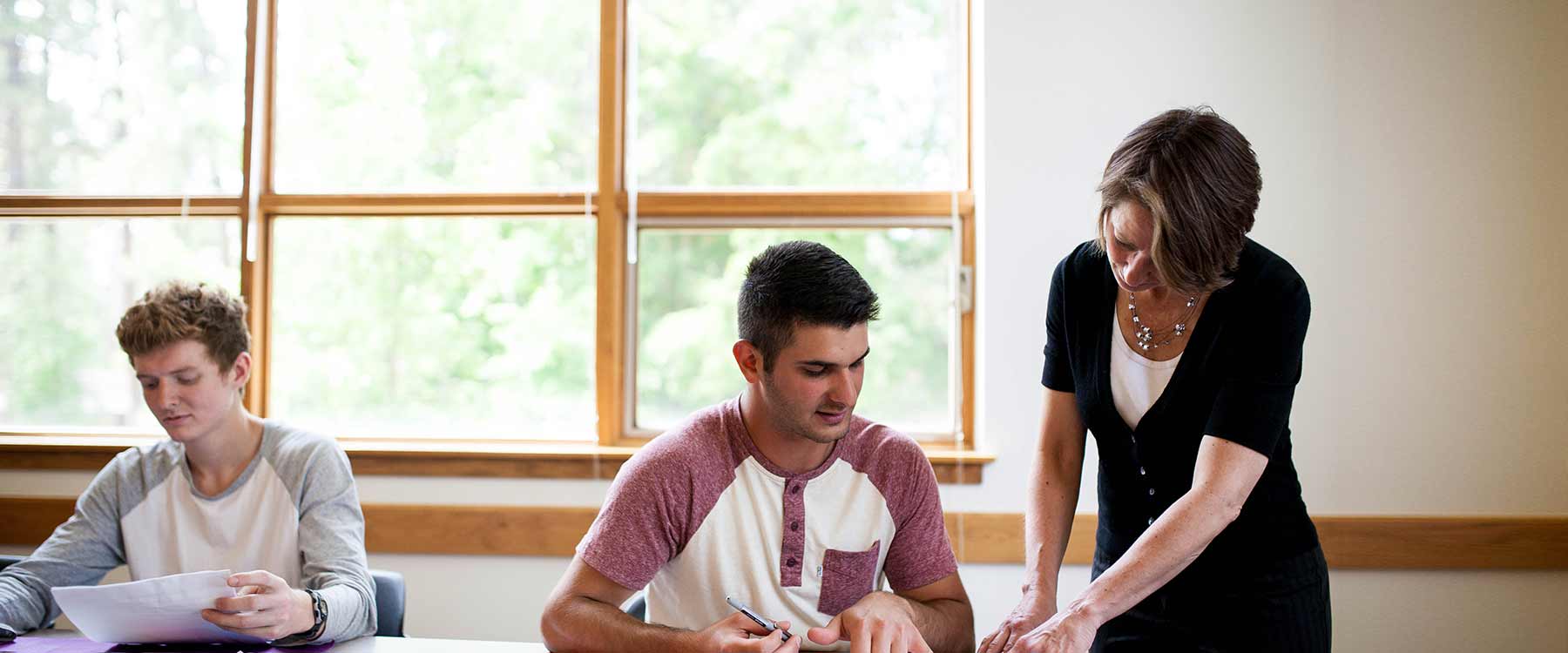 A professor stands and assists a student at a desk while another student works nearby near large windows.