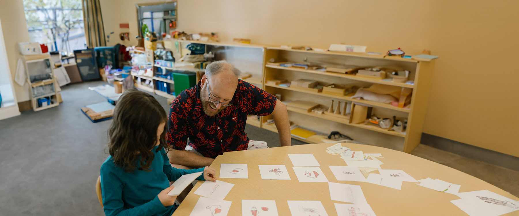 A teacher leans over a low table and flips through student artwork.