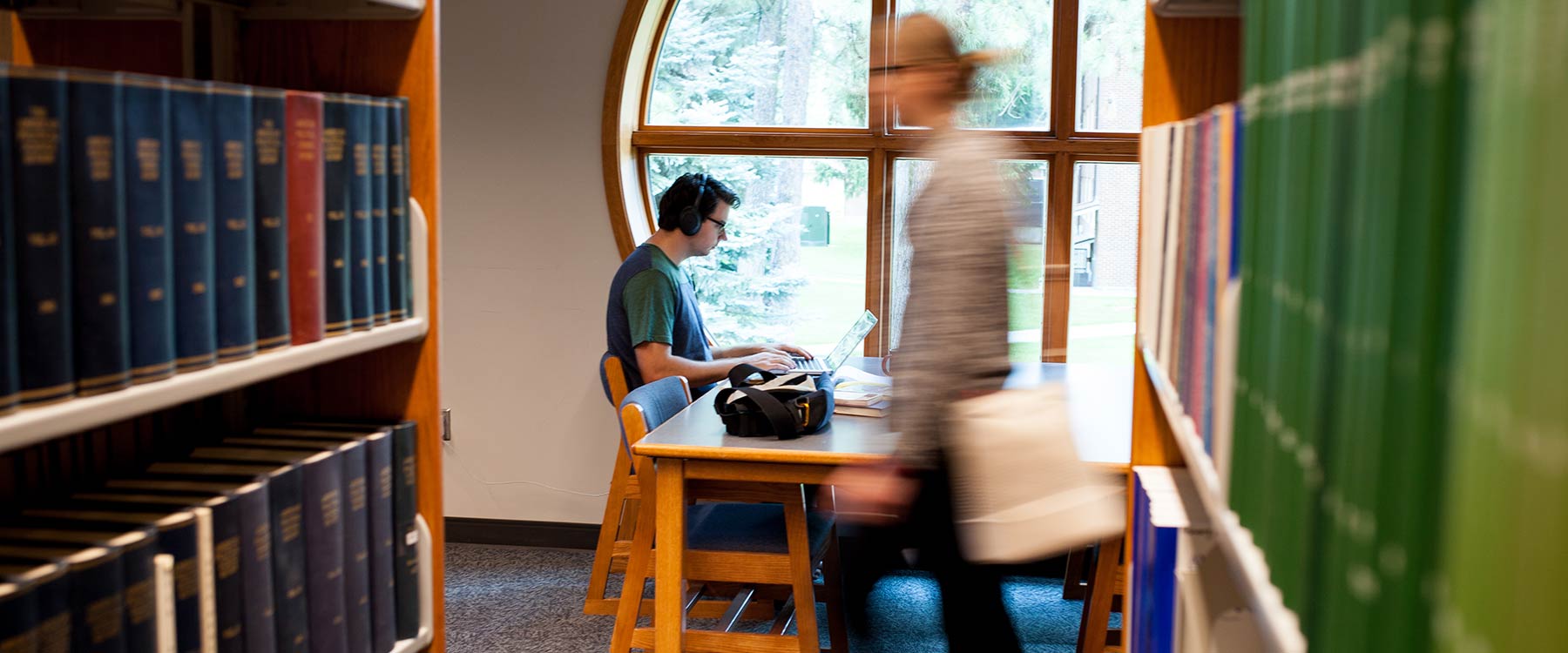 A student works on his laptop in the library, bookshelves surrounding him. Another student passes by.