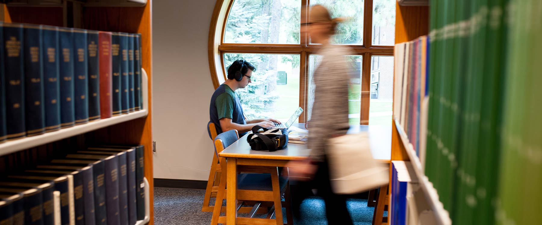 A student studies at a library table near a round window as another walks past.