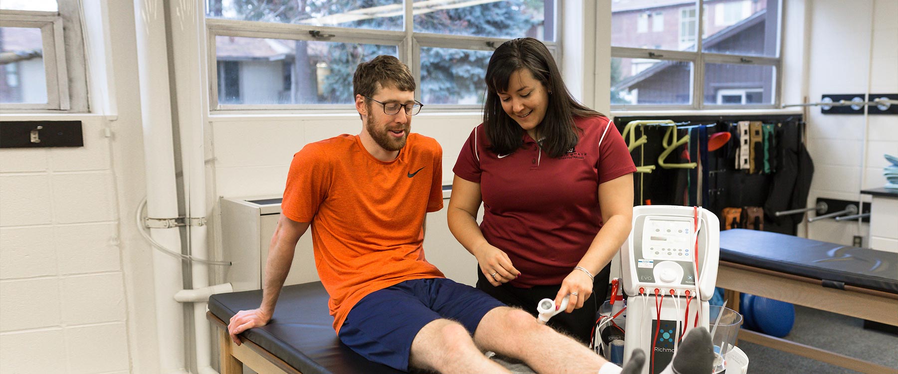 A student seated on an examination table receives treatment on his knee from another student in a clinic.