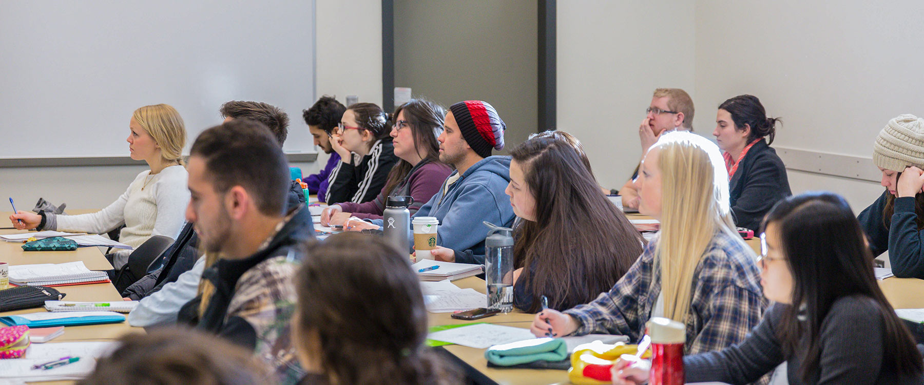 A group of students sit in rows in a full classroom listening and taking notes during a lecture.