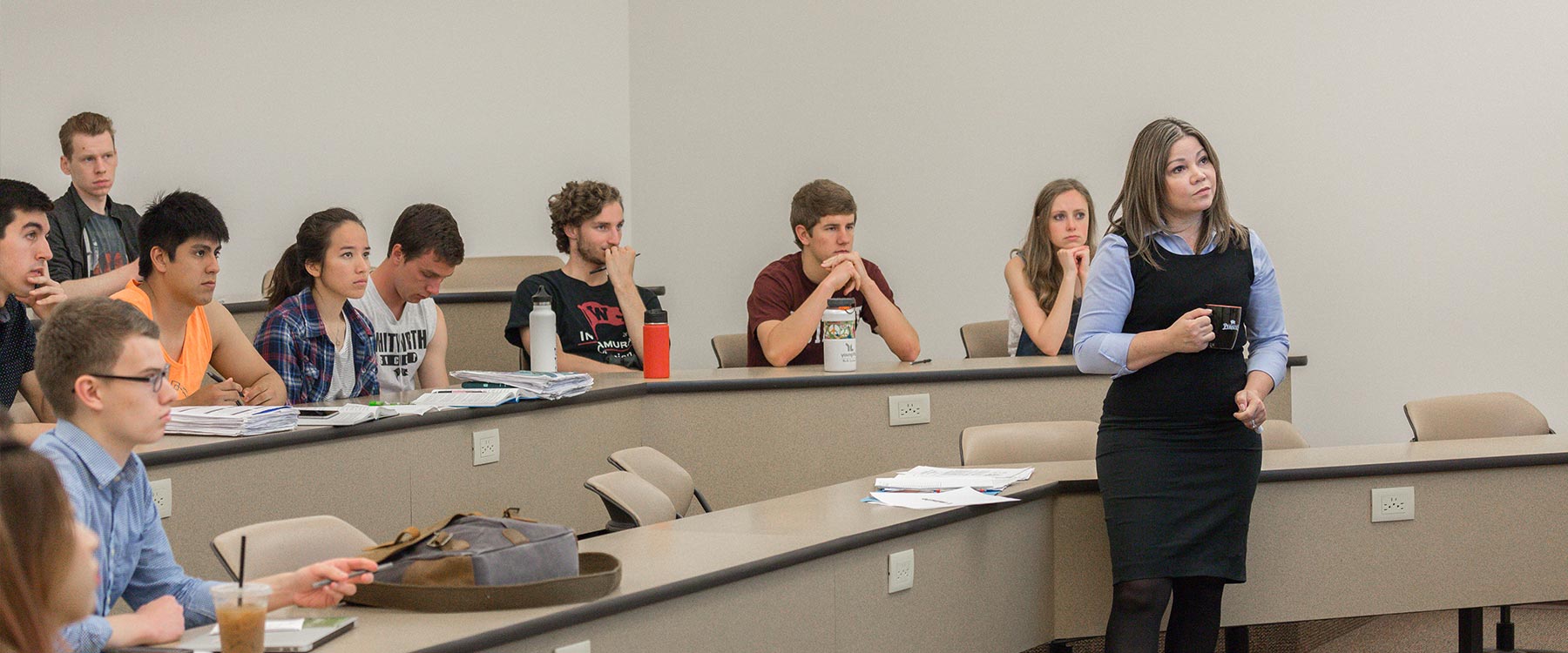 A professor stands in front of tiered rows of students holding a coffee cup. They all look toward the front of the classroom.