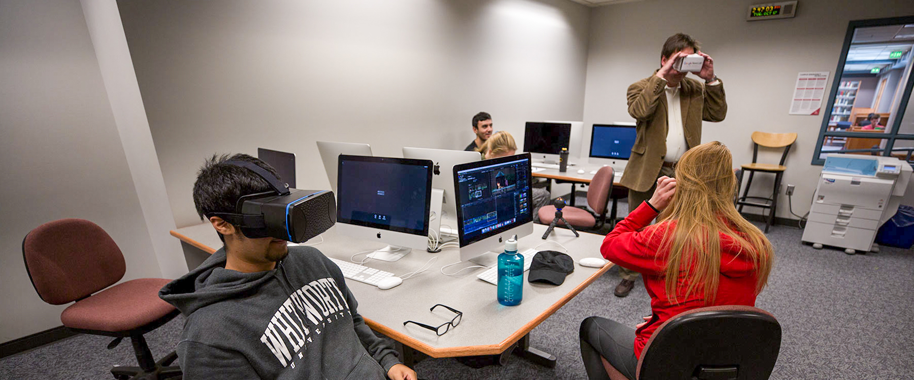 A student sits looking through virtual reality goggles while a professor and other students look through more sets of goggles.