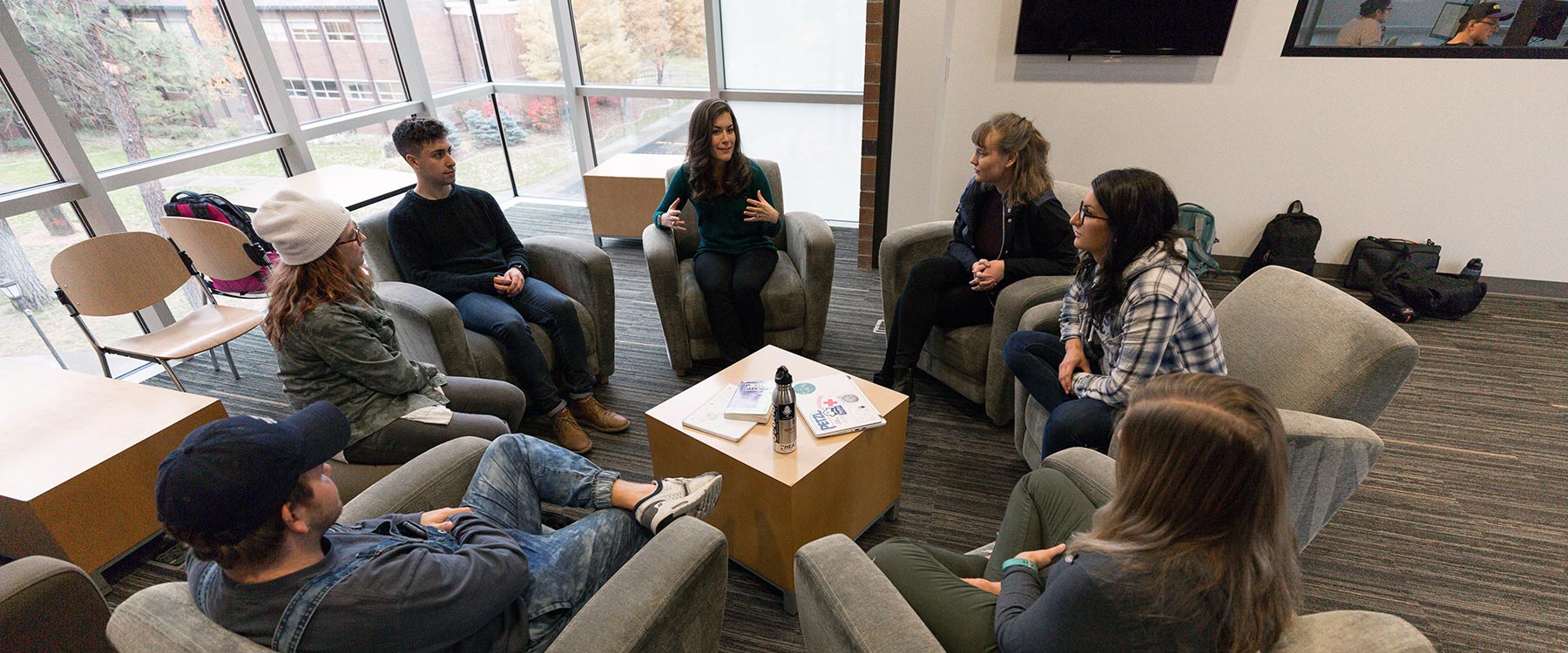 A professor and six students sit in a circle of armchairs, conversing. Nearby windows look out over campus.