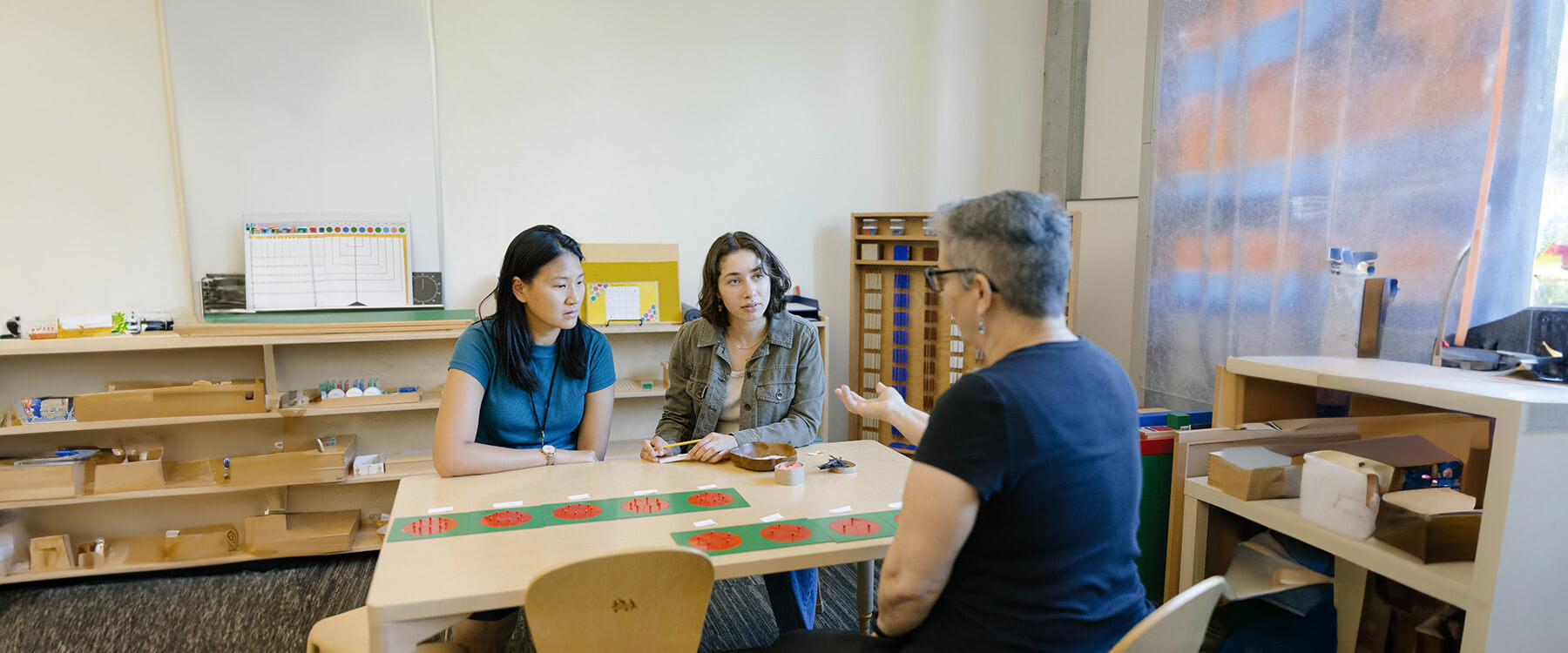 A teacher sits across from two students at a table discussing an elementary fractions project.