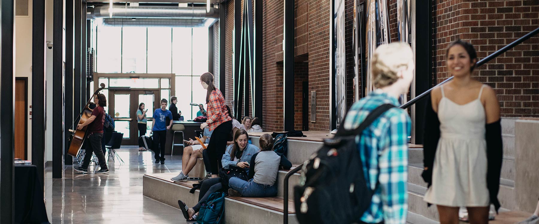 Students study and socialize in a bright, spacious corridor. Two are carrying large instruments.