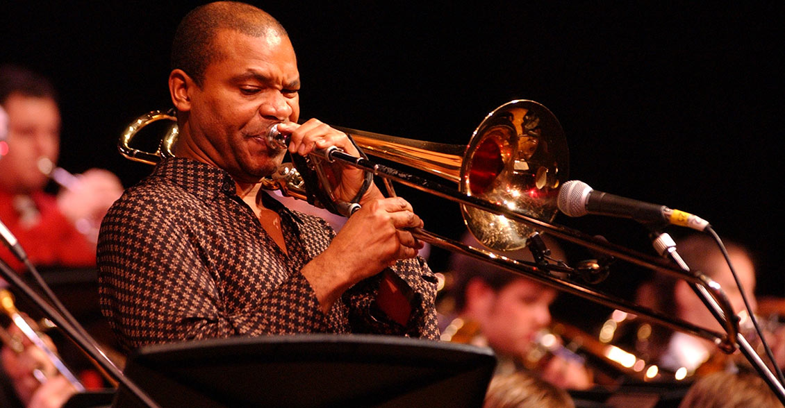 Robin Eubanks in a checkered shirt playing trombone into a microphone, under lights.