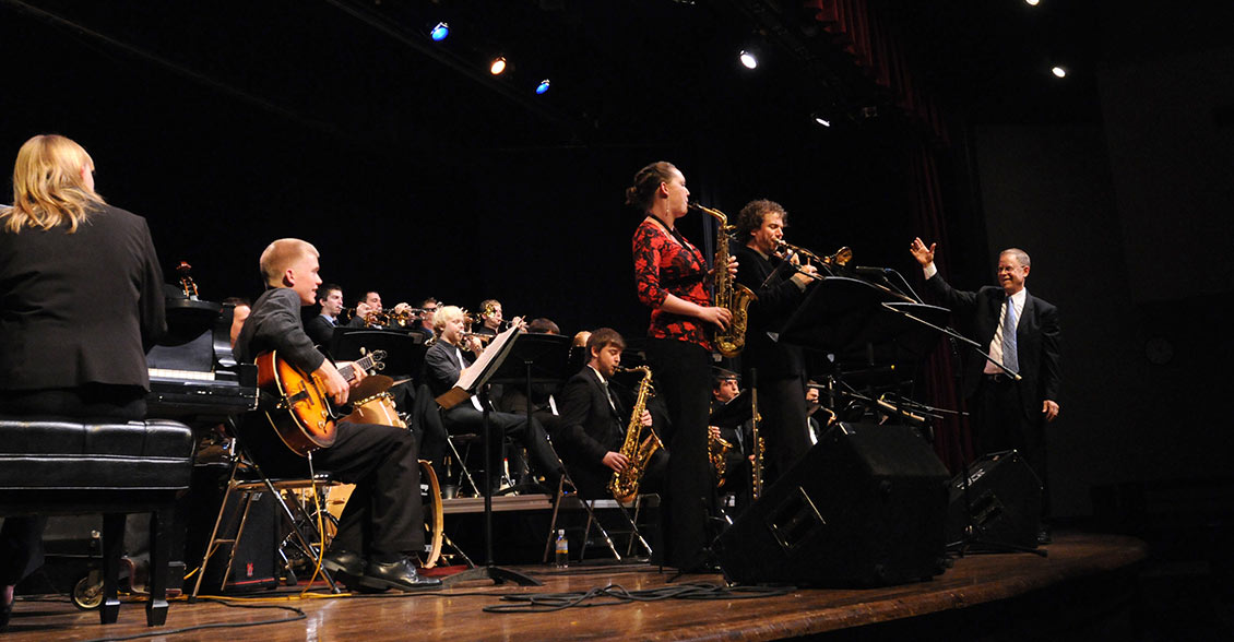 Two artists soloing while the band director stands to the side, smiling and gesturing towards them.