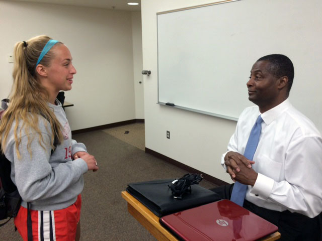 A student stands and talks to a professor who sits behind a podium in front of a whiteboard.