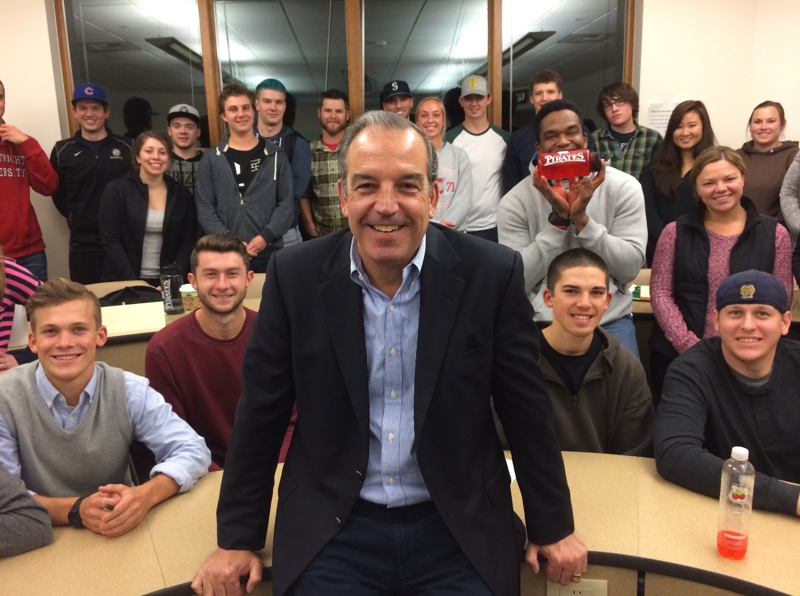 Augie Cruciotti sits in the forefront on the edge of a desk, smiling at the camera, with students smiling and posing in the background.