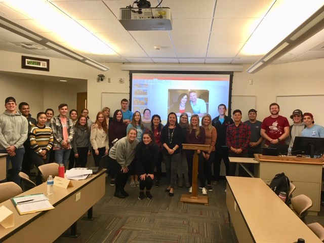 Daria Brown and students pose, smiling, in front of a projector screen in a classroom setting.