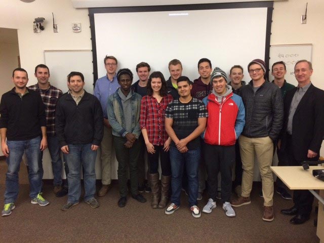 A professor and students pose, smiling, in front of a whiteboard in a classroom setting.