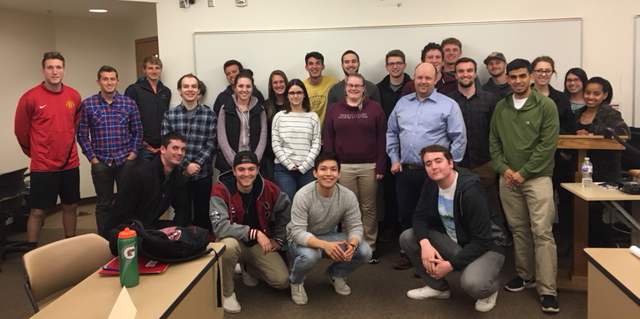Josh Hug and students pose, smiling, in front of a whiteboard in a classroom setting.