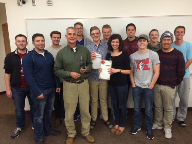 A professor and students pose, smiling, in front of a whiteboard in a classroom setting.