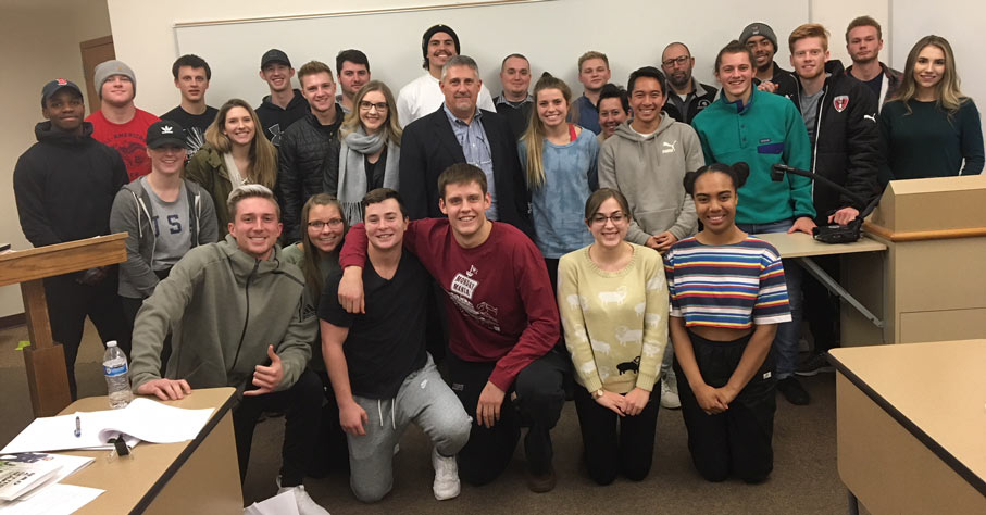 Stephen John and students pose, smiling, in front of a whiteboard in a classroom setting.