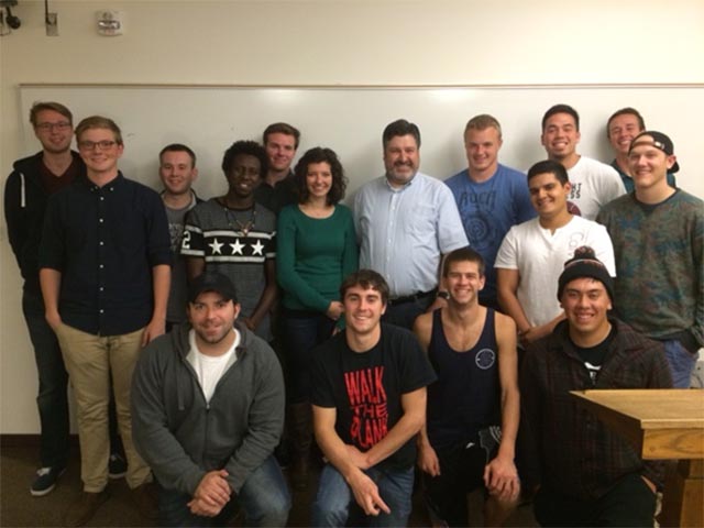 A professor and students pose, smiling, in front of a whiteboard in a classroom setting.