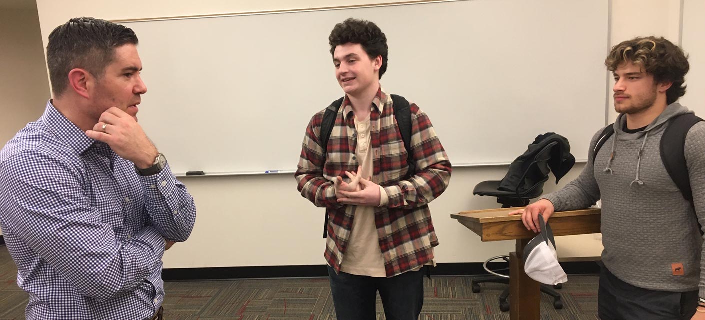 Two students and a professor engage in conversation in a classroom setting.