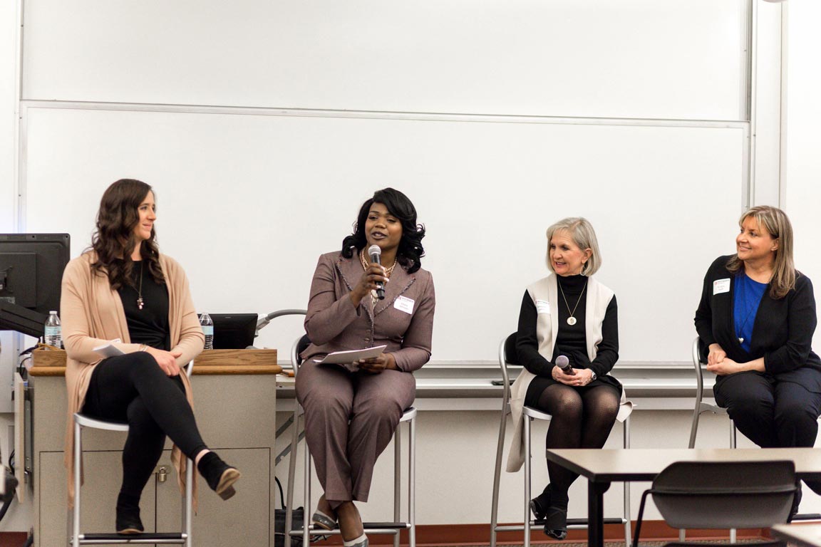 Four professionally dressed women sit in front of a classroom and one speaks into a microphone.