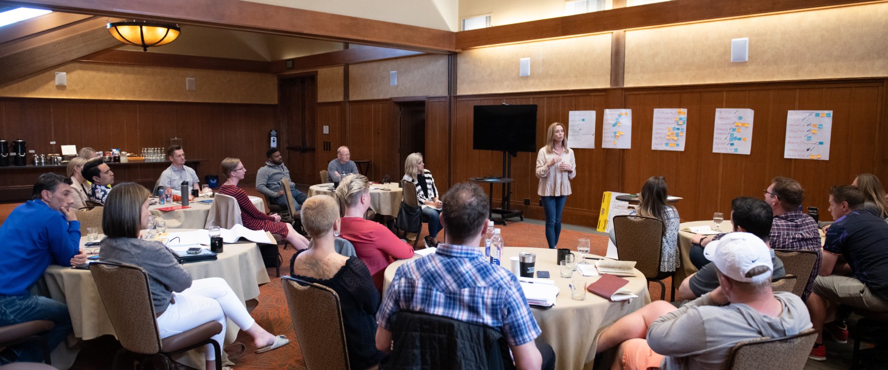 A presentor stands and presents to a group of people seated around tables in a conference room.