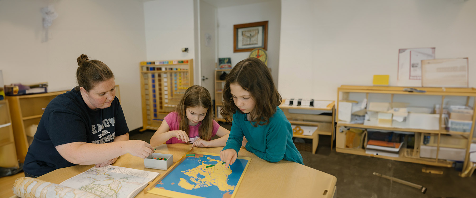 A teacher bends over a low table and flips through a stack of student art projects.