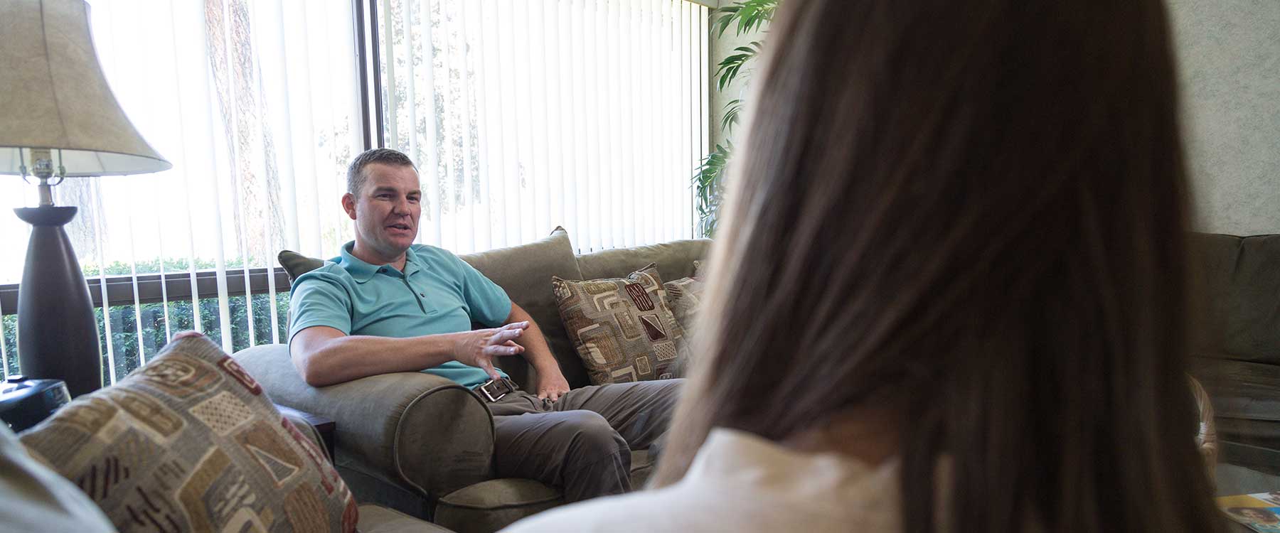One person, seen from behind, listens as another sits casually on a couch, talking.