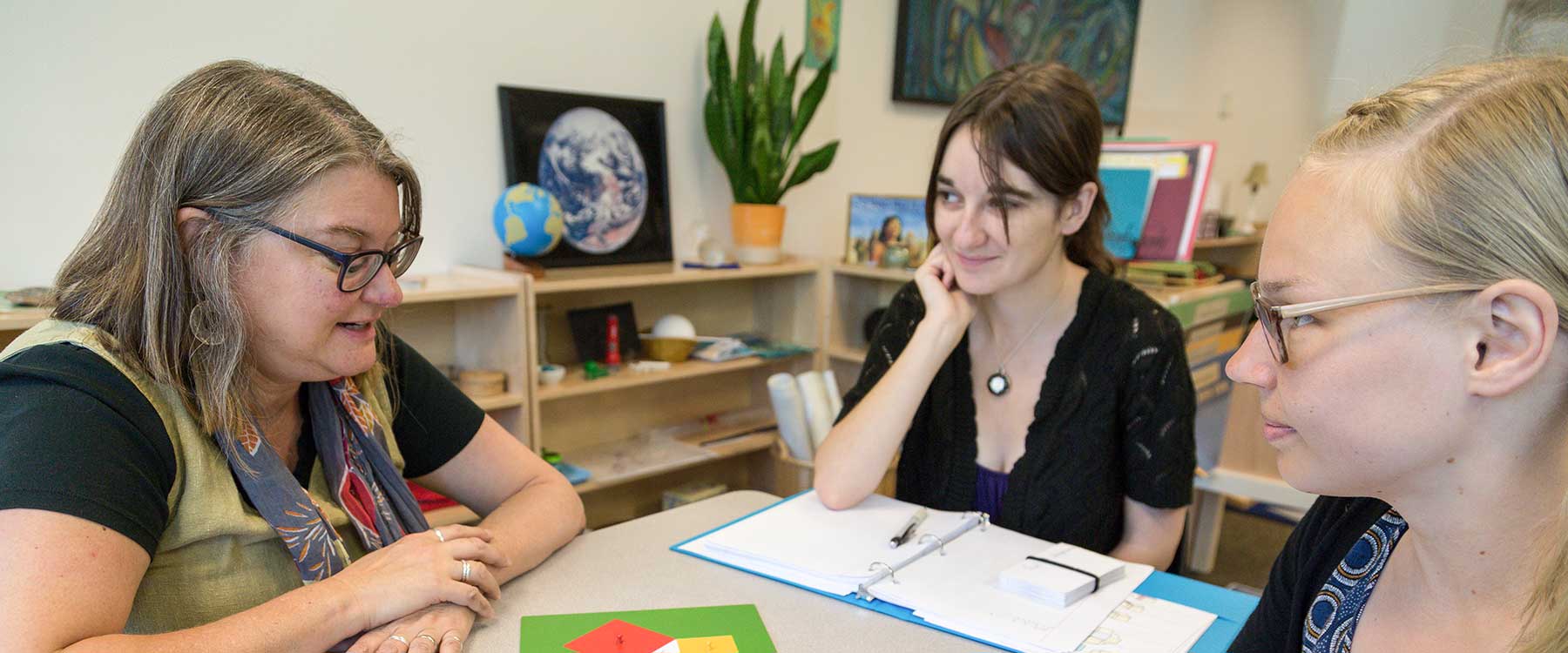 A teacher sits with two students at a small table. The students listen as the teacher talks.