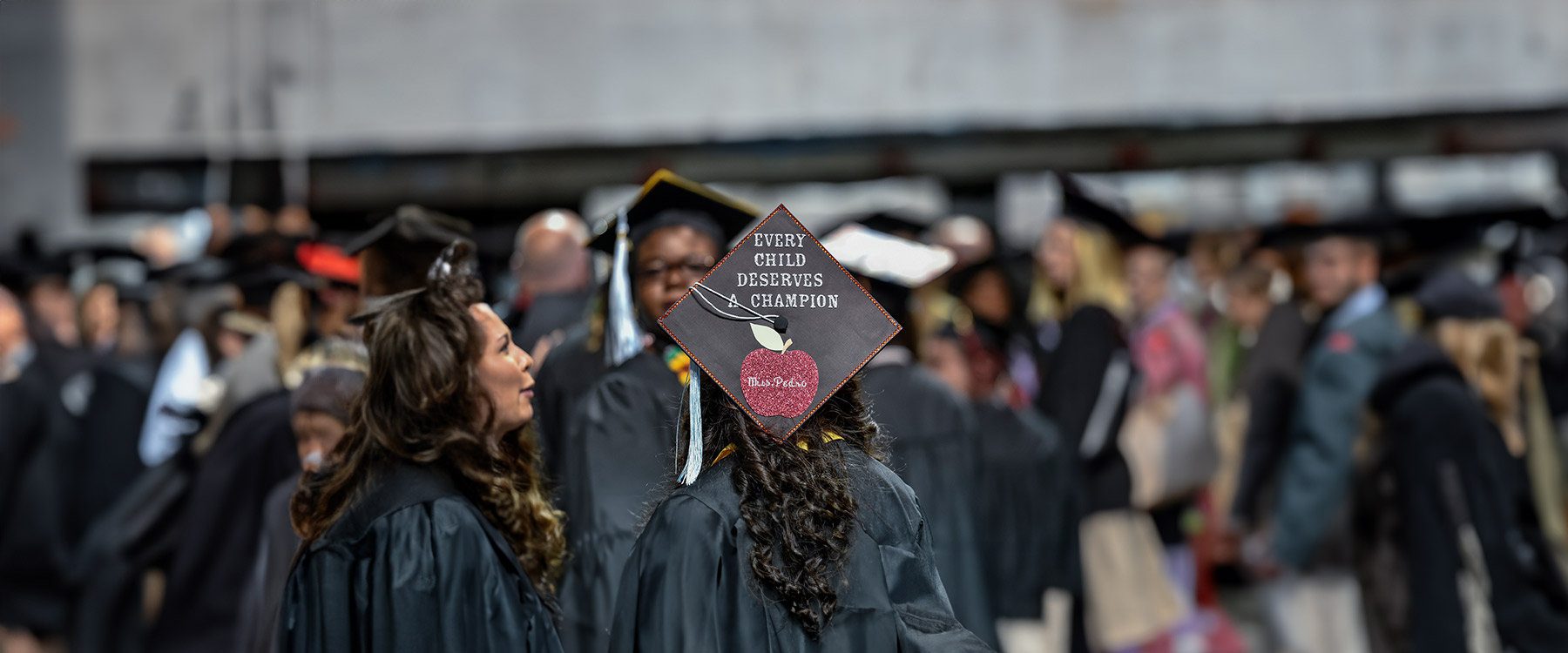 Three graduates in cap and gown stand together talking in a crowd, one with a cap that reads, 