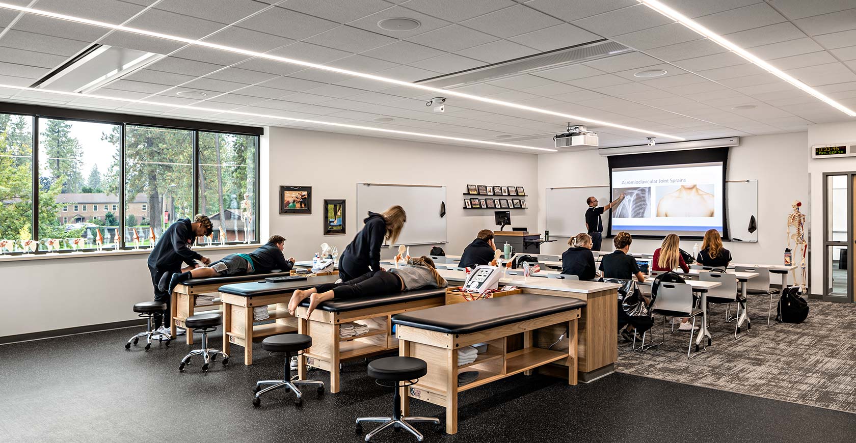 An instructor points to an anatomical figure on a screen while students observe, some seated and some at treatment tables.