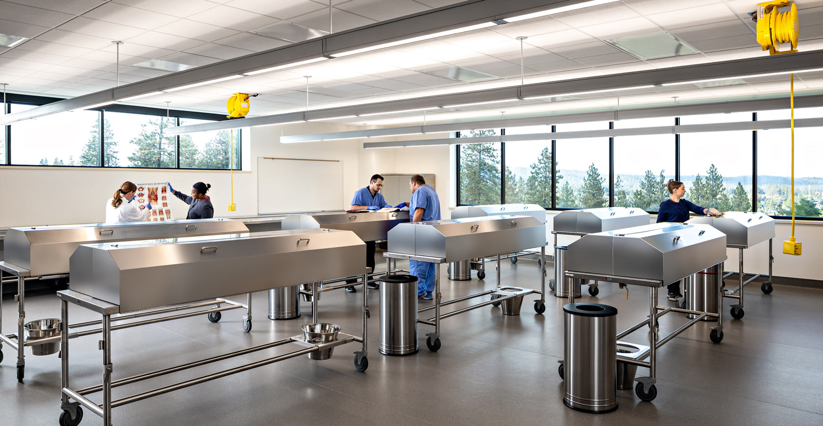 Students and professors study documentation within a cadaver lab with large windows overlooking a forested hill.