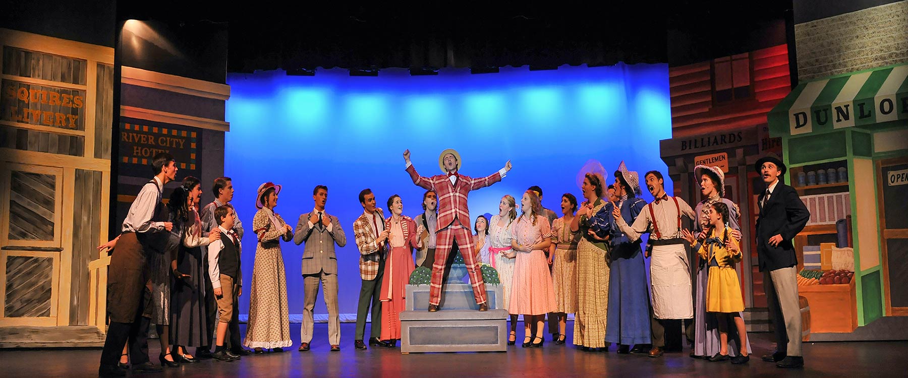 A theater cast in colorful costumes performs a lively musical number on stage, featuring a performer in the center.