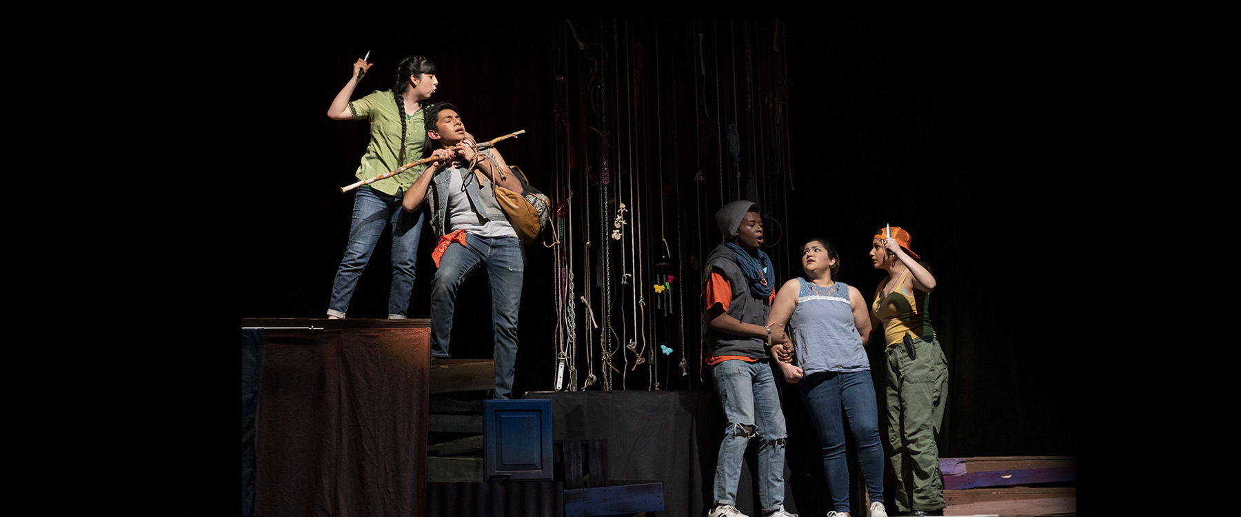 Five actors perform a dramatic scene on stage, two elevated on a platform while three stand below.